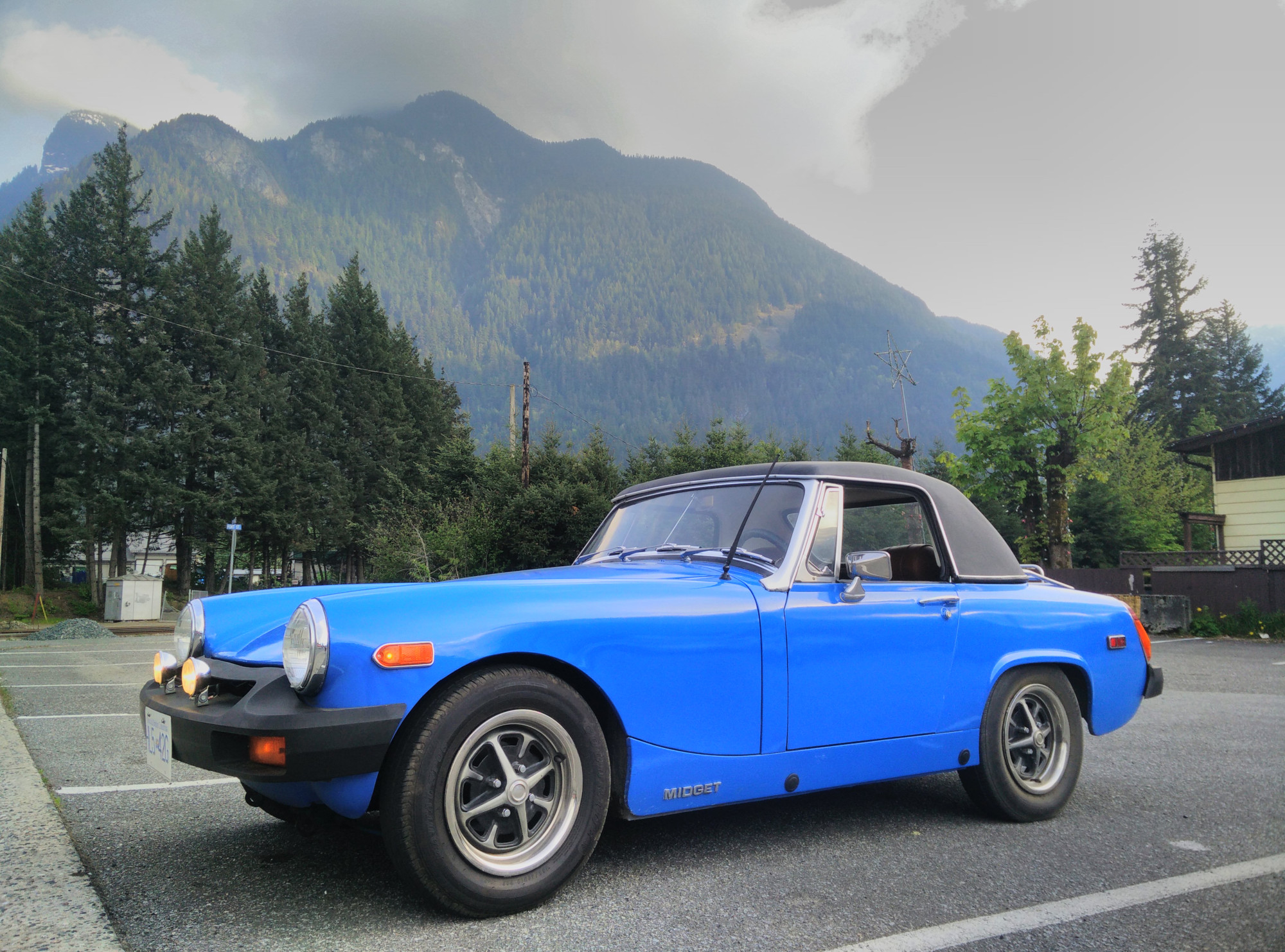 A very small blue convertible car from the 1970s is parked in an empty car park with a large, forested mountain and cloudy skies behind.