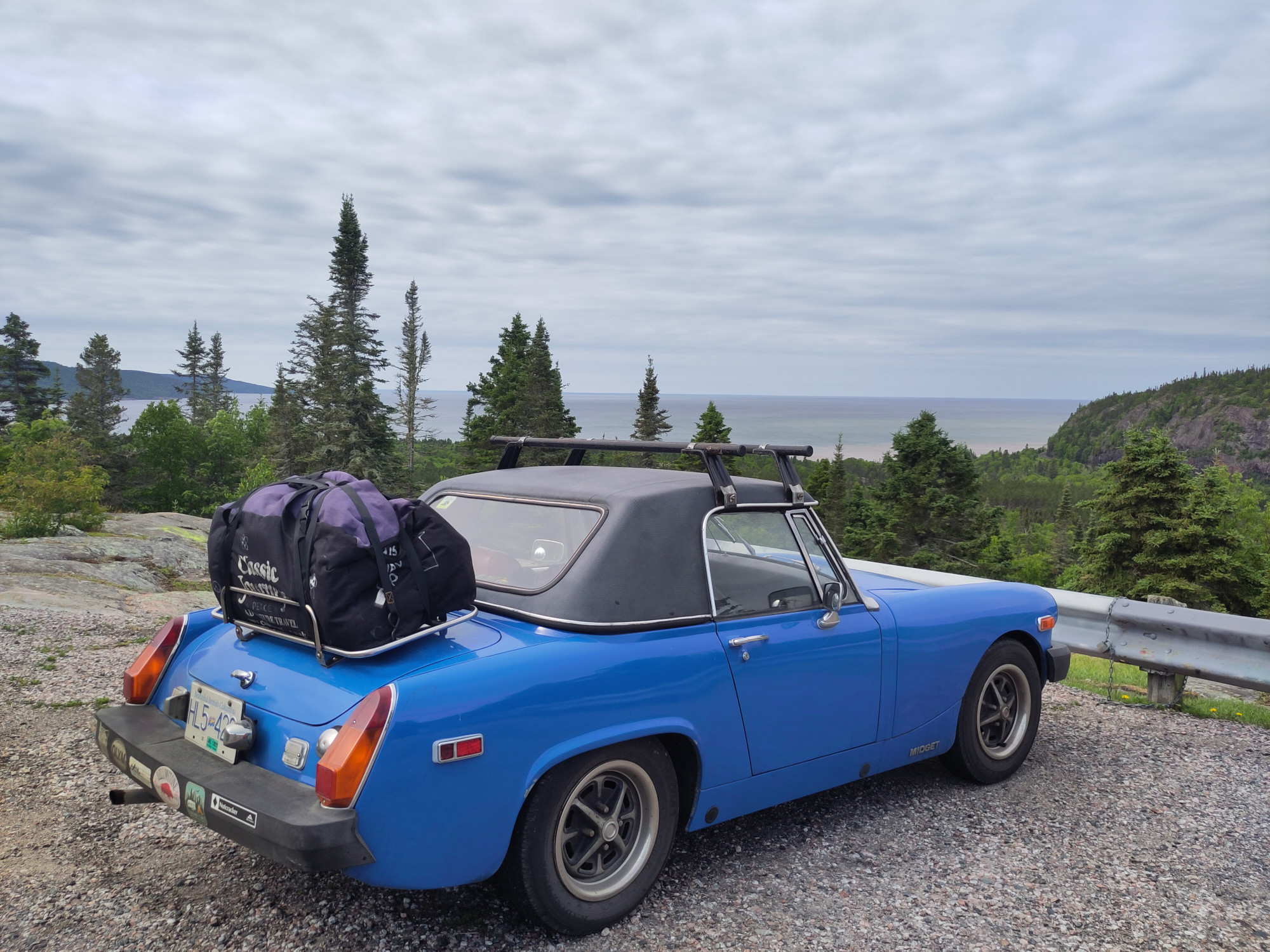 A small blue car from the 70s in a lay-by overlooking Lake Superior in the background. The car is loaded down with a large duffel bag on the rear luggage rack.