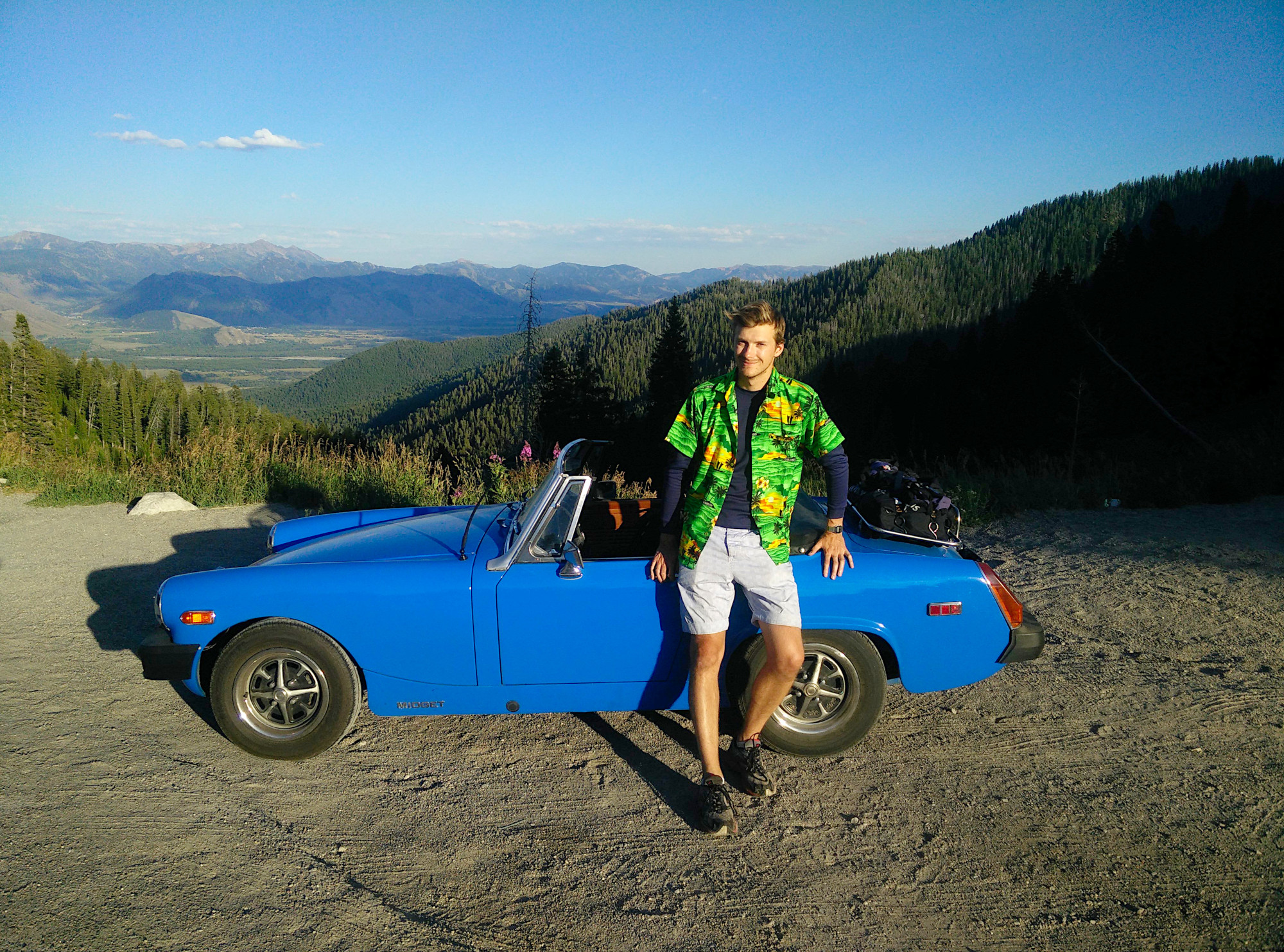 The same small blue car in a lay-by overlooking Teton Pass, with the author wearing light grey shorts and a horrible floral green shirt leaning on it.
