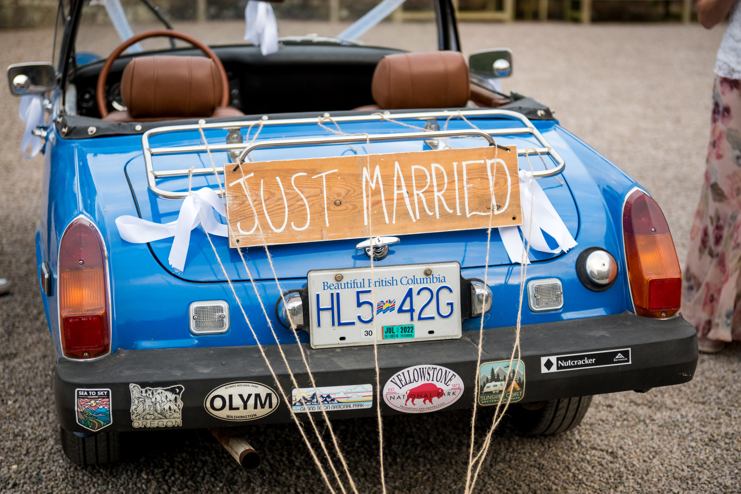 The rear view of a vintage blue convertible decorated with white ribbons tied to its chrome luggage rack. A rustic wooden sign reading 'Just Married' is attached above the license plate.