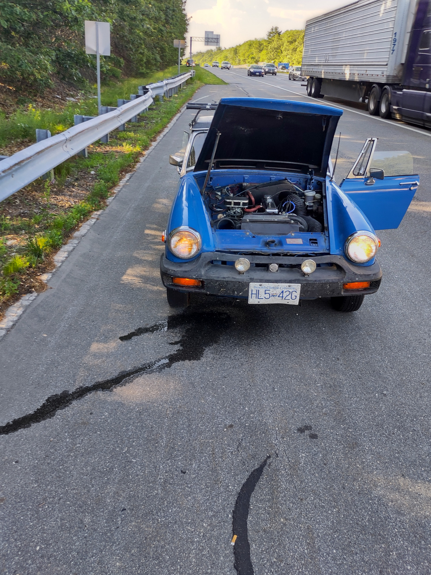 A small blue car broken down on a freeway hard shoulder with the hood up and coolant spilling all over the pavement.