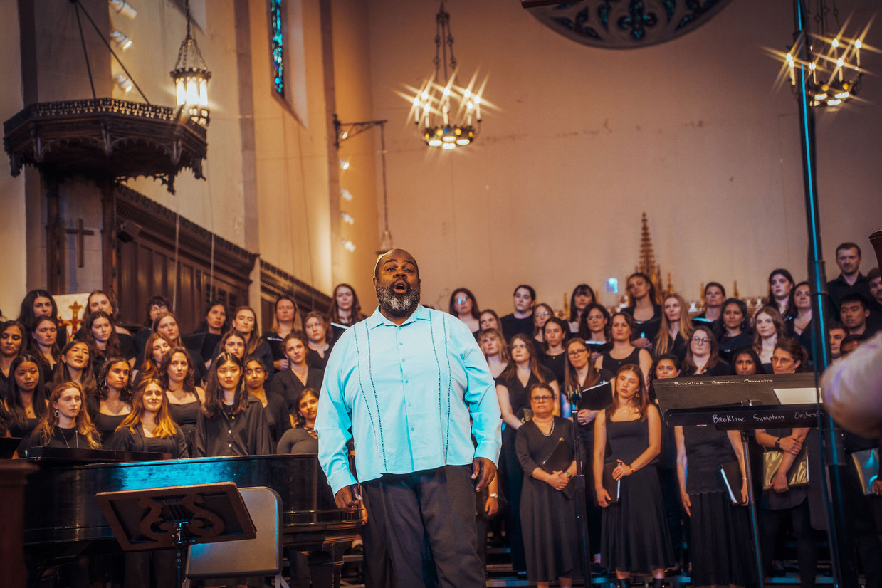 A man in a light blue button-down shirt stands center stage with his mouth open, singing passionately. Behind him, the Longwood Chorus—dozens of performers dressed in black—is arranged on risers inside a church-like hall with high ceilings and hanging chandeliers.