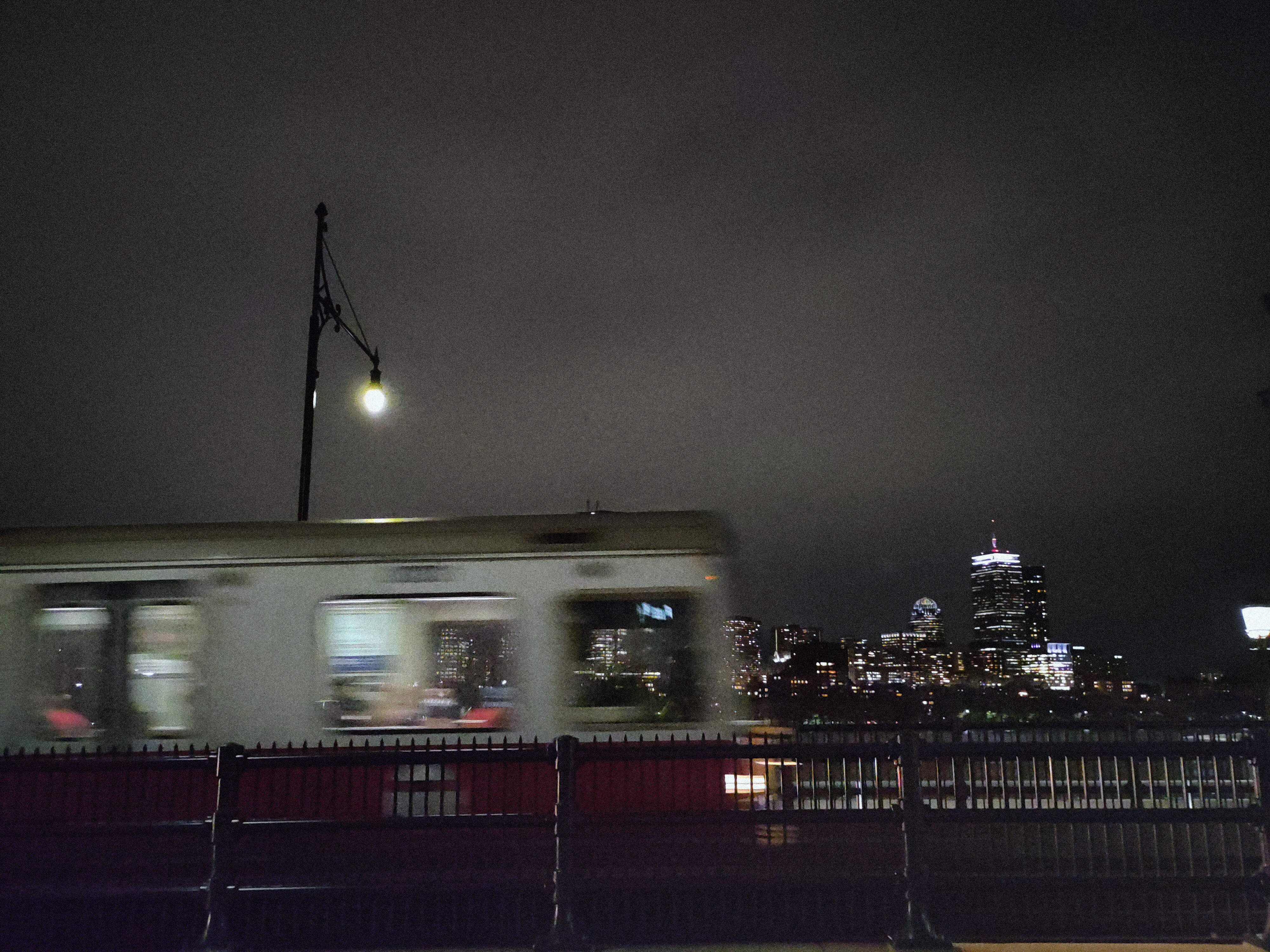 A view from a bridge of the Boston skyline is interrupted by a red line train crossing the frame in the foreground, slightly motion blurred.